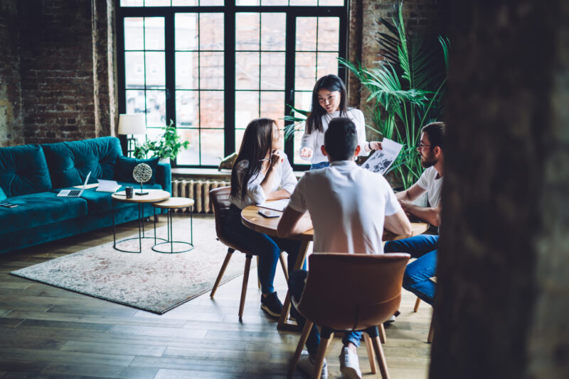 Positive young multiracial coworkers wearing blue jeans and white shirts gathering at table and sharing ideas while working together in creative coworking space